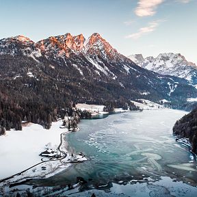 Verschneiter Bergsee umgeben von bewaldeten Bergen unter blauem Himmel. Die winterliche Landschaft strahlt Ruhe und natürliche Schönheit aus.