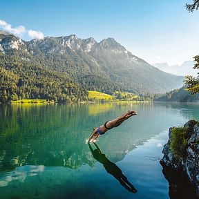 Person taucht in klaren Bergsee mit Wald und Bergen im Hintergrund. Sonniges Wetter, ruhige Atmosphäre, grüne Umgebung und malerische Landschaft.