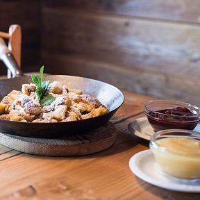 Ein Teller Kaiserschmarrn mit Puderzucker, serviert in einer Pfanne auf einem Holztisch. Dazu gibt es kleine Schälchen mit Apfelmus und Preiselbeermarmelade.