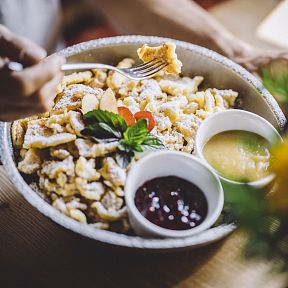 Ein Teller mit Kaiserschmarrn, bestäubt mit Puderzucker, serviert mit Apfelmus und Beerenkompott. Im Vordergrund unscharf ein paar Blumen.