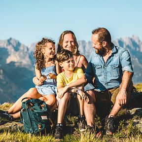 Eine fröhliche Familie sitzt auf einer Wiese vor beeindruckenden Bergen. Die Sonne scheint, und alle lächeln, während sie die Bergkulisse im Hintergrund genießen.