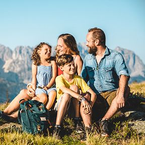 A family of four sits on a grassy hill with backpacks, enjoying the sunshine. Behind them is the breathtaking view of the Wilder Kaiser mountains.