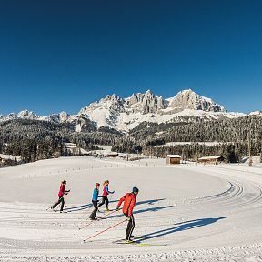 Menschen beim Langlaufen auf einer verschneiten Strecke vor einer Kulisse von schneebedeckten Bergen unter klarem, blauem Himmel.