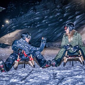 Two people in winter gear sit on sleds, smiling and enjoying a night sledding adventure on a snow-covered path surrounded by trees at Wilder Kaiser.