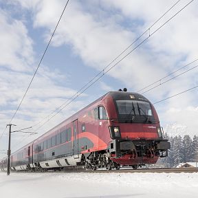 A red and blue train traveling through a snowy landscape near the Wilder Kaiser mountains under a partly cloudy sky.