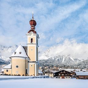 Snowy village with a church near the Wilder Kaiser mountains under a blue sky.