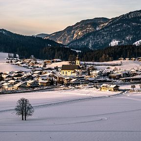 A snow-covered village nestled in the Wilder Kaiser region, with mountains in the background and warm sunlight illuminating the landscape.