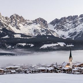 Snowy Wilder Kaiser mountains rise above a fog-covered village with a church spire. The landscape is blanketed in white, offering a serene winter view.