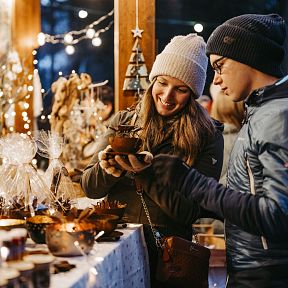 Ein Paar erkundet einen festlichen Weihnachtsmarktstand. Sie tragen warme Winterkleidung und betrachten kunstvoll gefertigte Geschenke bei festlicher Beleuchtung.