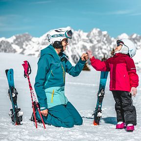 Parent and child in ski attire enjoying a moment on the snowy slopes of the Wilder Kaiser, with impressive alpine peaks in the background.