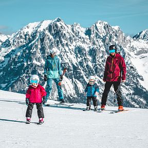 Eine Familie mit zwei Kindern steht in bunter Skibekleidung auf einer schneebedeckten Piste, umgeben von majestätischen, verschneiten Berggipfeln.