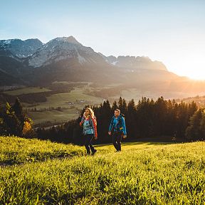 Zwei Wanderer gehen über eine grüne Wiese bei Sonnenuntergang in einer bergigen Landschaft. Im Hintergrund erheben sich beeindruckende Berggipfel.