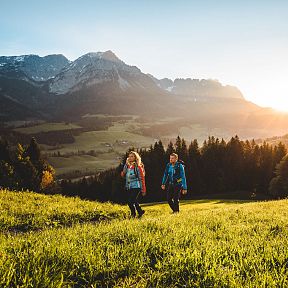 Two hikers on a grassy meadow in Wilder Kaiser, with the sun rising over majestic mountains in the background. A serene and stunning outdoor scene.