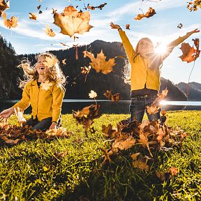Two children playfully throwing autumn leaves in the air on a grassy field with mountains and a clear sky in the background.