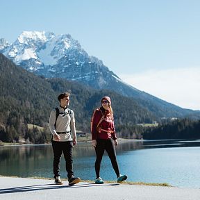 Zwei Menschen wandern an einem See, umgeben von bewaldeten Bergen und schneebedeckten Gipfeln unter blauem Himmel. Ein idyllisches Naturmotiv.
