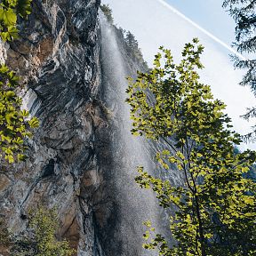 Ein Wasserfall fließt aus einer steilen Felswand inmitten einer bewaldeten Berglandschaft, umgeben von grünem Laub und blauem Himmel im Hintergrund.