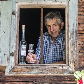 Ein älterer Mann in einem karierten Hemd steht an einem Holzfenster und hält ein Glas Schnaps neben einer Schnapsflasche.