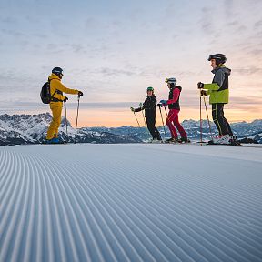 Vier Skifahrer stehen auf einer präparierten Skipiste im Vordergrund, während im Hintergrund Berge und ein Sonnenuntergang zu sehen sind.