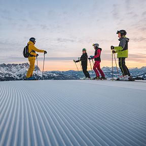 Vier Skifahrer stehen auf einer präparierten Skipiste im Vordergrund, während im Hintergrund Berge und ein Sonnenuntergang zu sehen sind.