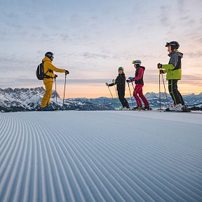 A group of skiers stand on a groomed slope at sunrise, with mountain peaks in the background and colorful ski gear creating a vibrant scene.