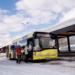 A bus parked at a snow-covered station in the Wilder Kaiser region, with people in winter gear preparing to board. Snow-capped mountains can be seen in the background.