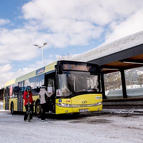 A bus parked at a snow-covered station in the Wilder Kaiser region, with people in winter gear preparing to board. Snow-capped mountains can be seen in the background.