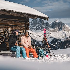 Zwei Menschen sitzen in Skikleidung vor einer verschneiten Berghütte. Sie genießen die Winterlandschaft mit schneebedeckten Bergen im Hintergrund.