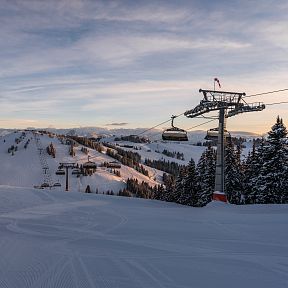 Snowy ski slopes with chairlift and distant mountain range under a partly cloudy sky, during sunset or sunrise. Coniferous trees line the landscape.