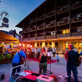 Eine Band spielt bei einem Straßenfest vor einem traditionellen Gebäude, während das Publikum bei abendlicher Stimmung auf der Terrasse sitzt.