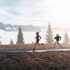 Two people running on a path in front of the Wilder Kaiser mountains, with mist and trees creating a serene, alpine landscape.