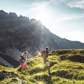 Zwei Wanderer gehen über eine grüne Bergwiese, im Hintergrund ragt ein imposantes Bergmassiv empor. Die Sonne scheint über den felsigen Gipfeln.