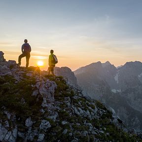 Zwei Wanderer stehen auf einem felsigen Gipfel und schauen in einen farbenfrohen Sonnenuntergang über einen Bergkamm. Der Himmel ist klar und friedlich.