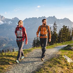 Zwei Menschen wandern auf einem Pfad mit Blick auf das beeindruckende Bergmassiv des Wilden Kaisers in Tirol. Die Sonne scheint, der Himmel ist blau.