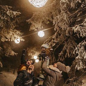 Familie genießt den Winter in einem verschneiten Wald mit Lichterketten. Ein Kind wird von einem Erwachsenen hochgehoben, alle sind warm angezogen und lächeln.
