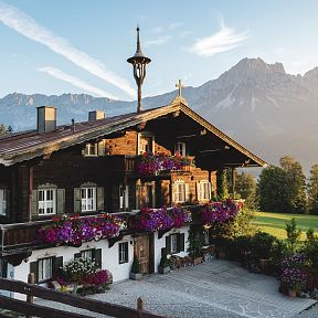 Traditionelles Tiroler Holzhaus mit blühenden Geranien, im Hintergrund das beeindruckende Bergmassiv des Wilden Kaisers bei Sonnenaufgang.
