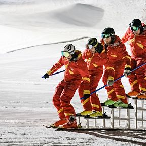 A synchronized ski team in bright red uniforms performs on a snowy slope in the Wilder Kaiser region, demonstrating skill and teamwork under clear blue skies.