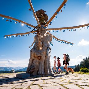 Drei Wanderer und ein Hund gehen an einer einzigartigen, skulpturartigen Holzinstallation vorbei, umgeben von Bergen und Wald unter einem klaren blauen Himmel.