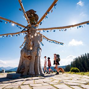 Drei Wanderer und ein Hund gehen an einer einzigartigen, skulpturartigen Holzinstallation vorbei, umgeben von Bergen und Wald unter einem klaren blauen Himmel.