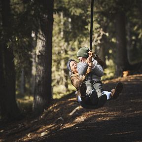 Zwei Menschen schwingen auf einer Seilbahn im Wald. Sie tragen warme Kleidung, während sie über den mit Laub bedeckten Waldboden gleiten. Sonnenlicht strahlt durch die Bäume.