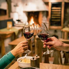 Zwei Personen stoßen mit Weingläsern an einem gemütlichen Holz-Tisch vor einem Kamin an, im Hintergrund ein warmes, einladendes Ambiente.