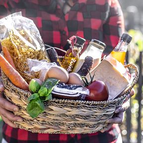 A person holding a wicker basket filled with fresh groceries including pasta, a carrot, eggs, cheese, a tomato, and a basil plant.