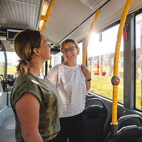 Zwei Frauen unterhalten sich in einem sonnendurchfluteten Bus. Die Landschaft draußen ist grün und ländlich. Beide lachen und scheinen die Fahrt zu genießen.
