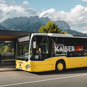 A yellow bus at a stop with people walking nearby, set against the backdrop of the Wilder Kaiser mountains and a partly cloudy sky.