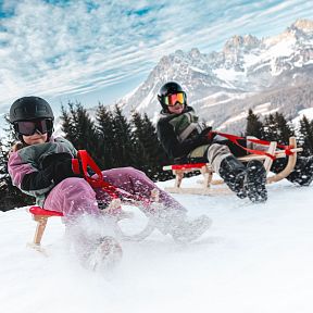 Two people sledding down a snowy hill with the majestic Wilder Kaiser mountains in the background, wearing winter gear and helmets.