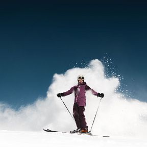 Eine Person in Skiausrüstung fährt dynamisch einen verschneiten Hang hinunter, umgeben von aufwirbelndem Schnee vor klarem, blauem Himmel.