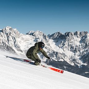 Ein Skifahrer in voller Fahrt auf einer schneebedeckten Piste mit den beeindruckenden Gipfeln des Wilder Kaiser Gebirges im Hintergrund, unter blauem Himmel.