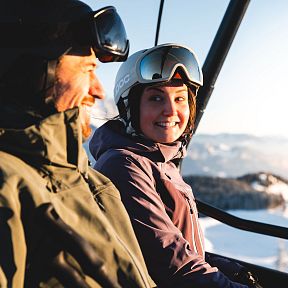 A man and woman smiling in a ski lift with snow-covered mountains and clear skies at the Wilder Kaiser in winter.