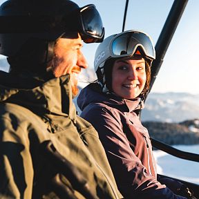A man and woman smiling in a ski lift with snow-covered mountains and clear skies at the Wilder Kaiser in winter.