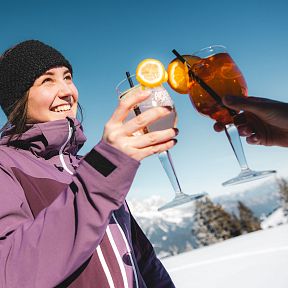 Frau in Winterjacke stößt mit anderen Personen mit Cocktails im Schnee an, vor blauem Himmel und Bergkulisse.