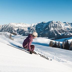 Eine Person fährt auf Skiern eine schneebedeckte Piste hinunter, umgeben von einem beeindruckenden Bergpanorama unter klarem, blauem Himmel.